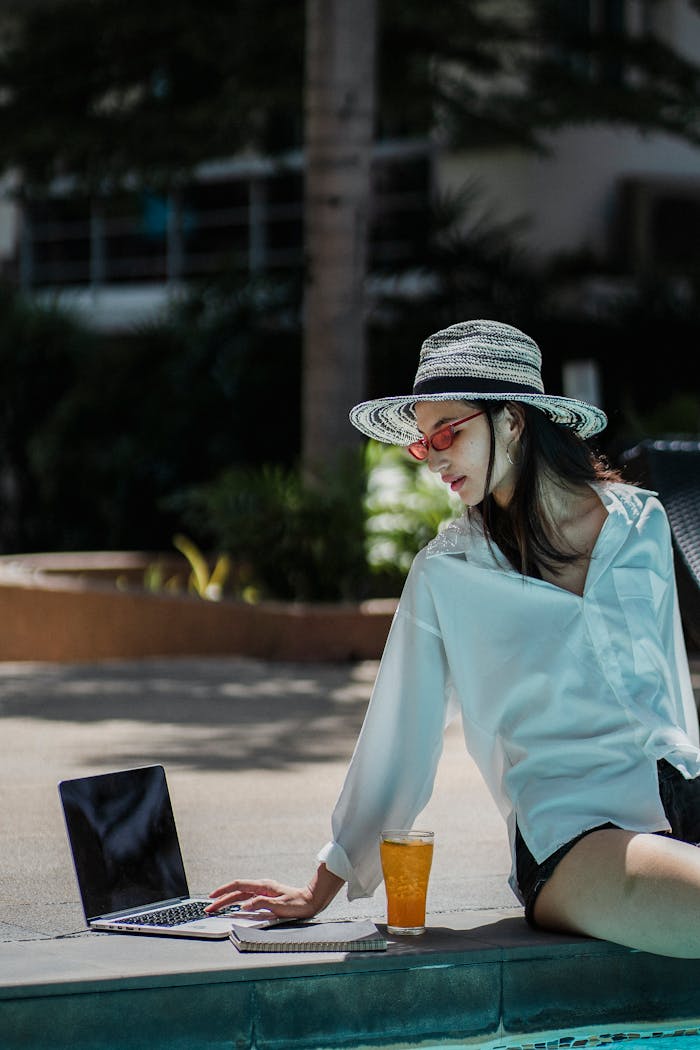 Young woman in straw hat working on a laptop by the poolside. Summer vibe.