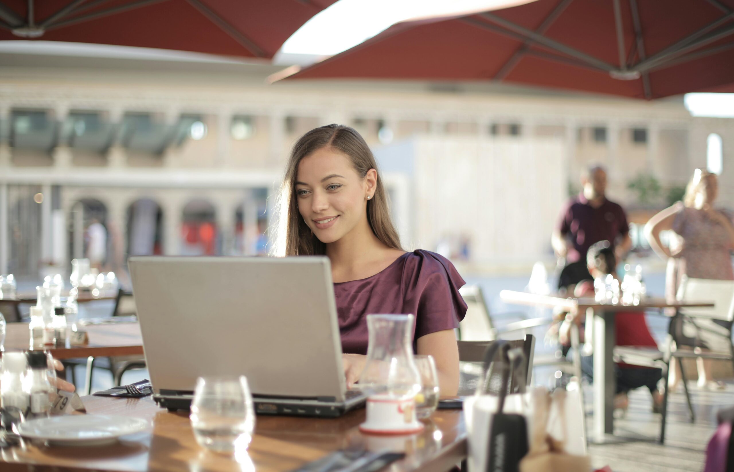 A woman sitting at an outdoor cafe, smiling and working on a laptop on a sunny day.