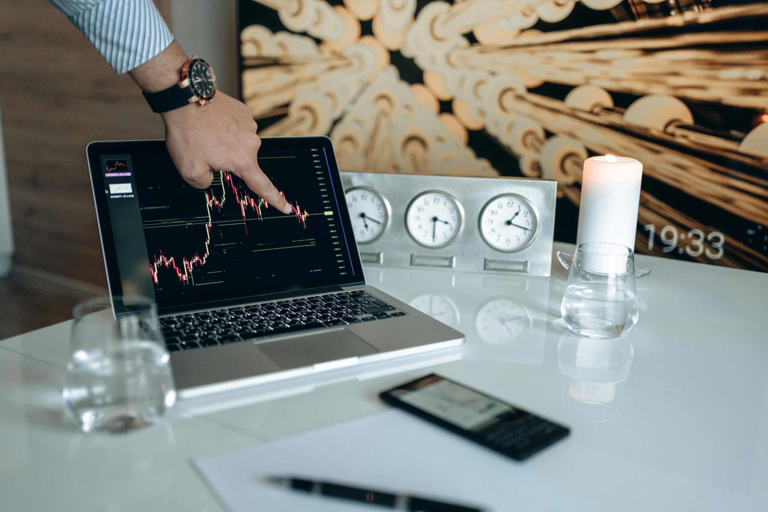 Hand pointing at market graph on laptop screen in modern office with clocks and candle.